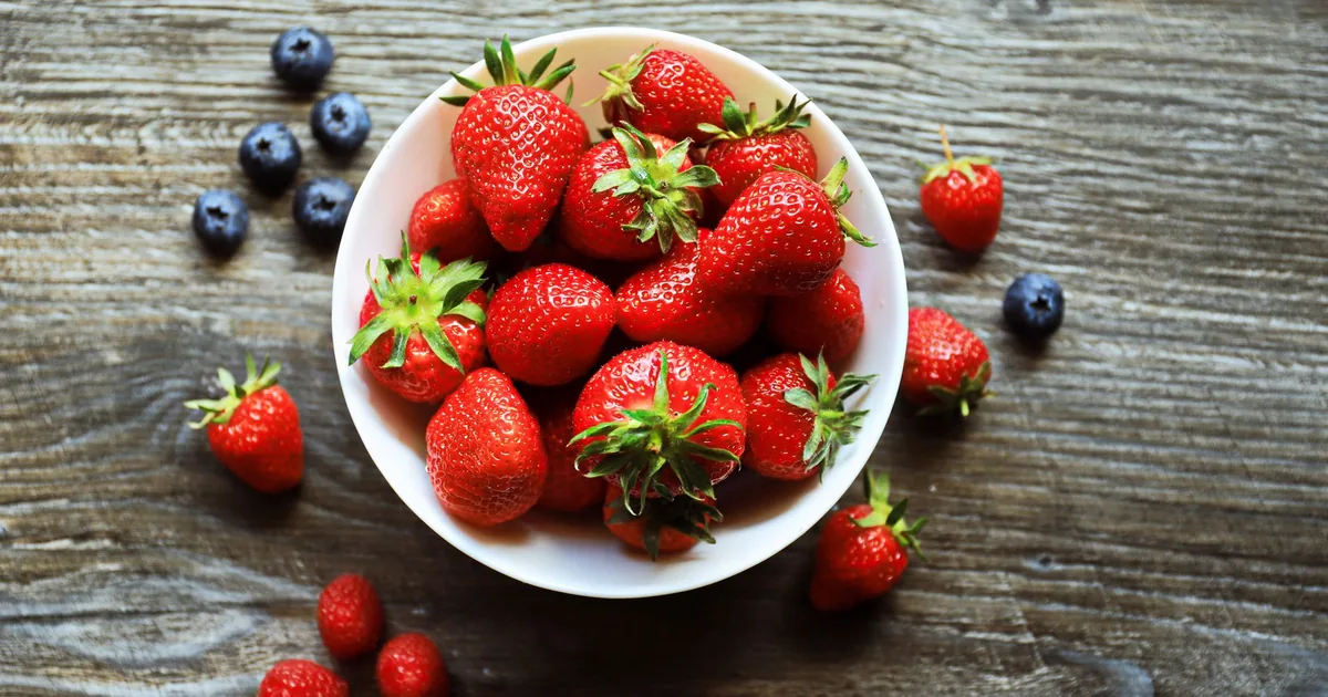 A bowl of fresh strawberries with blueberries and raspberries on a rustic wooden table, shot from above.