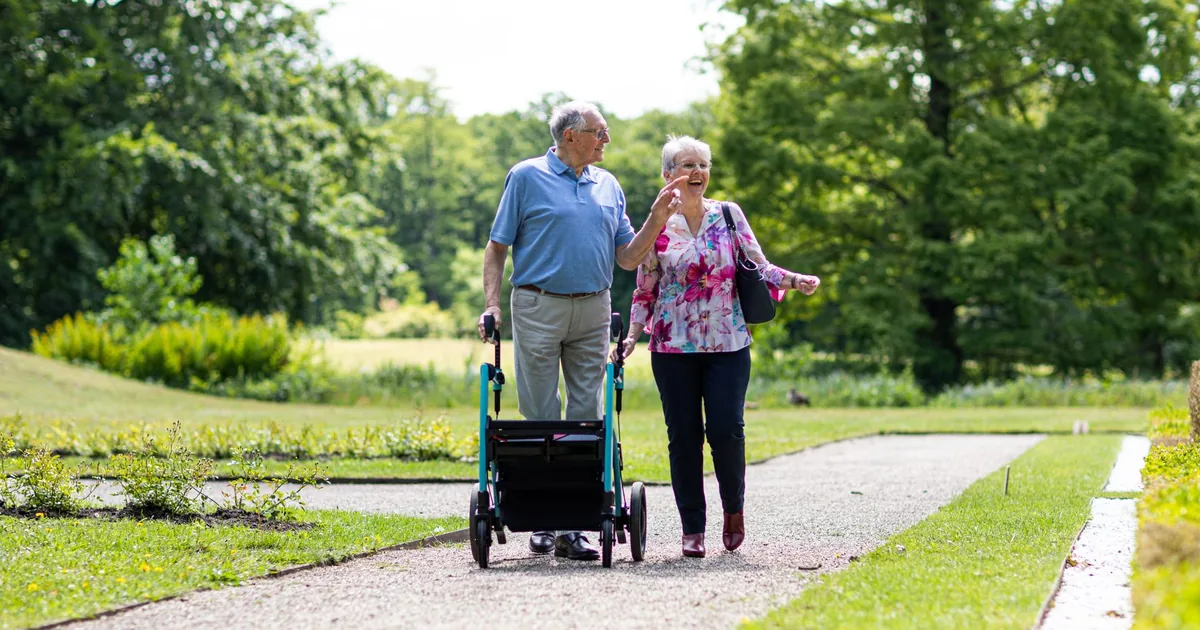 Senior couple walking in a park with a walker, enjoying a sunny day outdoors.