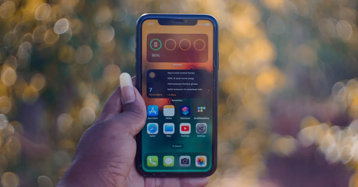 A hand holding a smartphone showing home screen apps, outdoors with bokeh background.