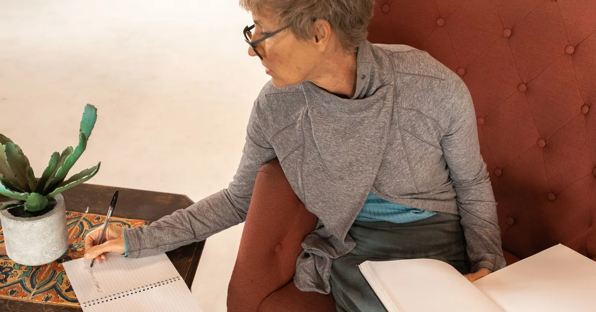 Elderly woman writing notes with a notebook while seated comfortably indoors.
