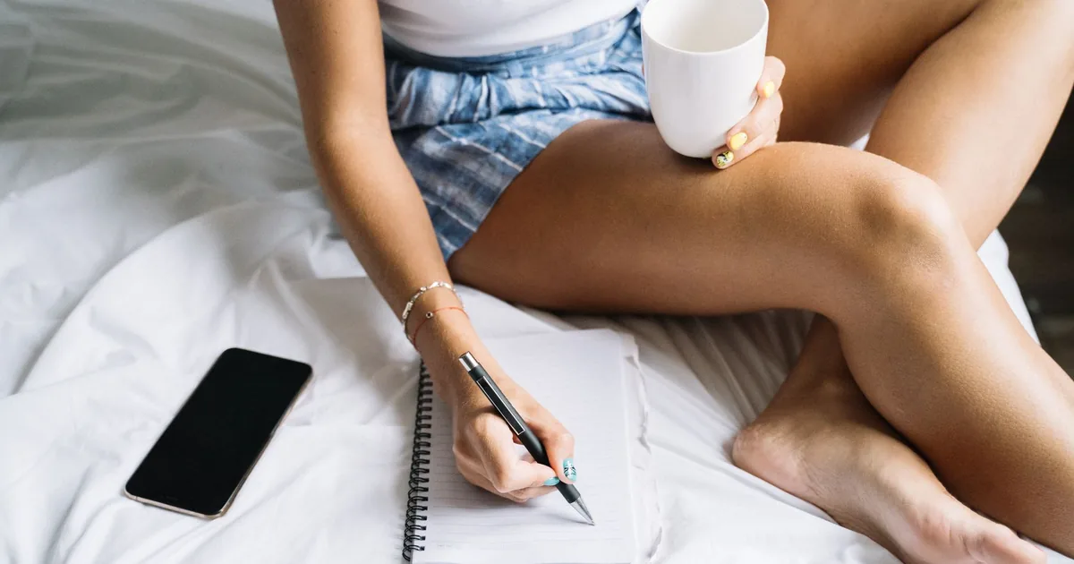 A woman holding a coffee cup while writing in a notebook, resting in bed.
