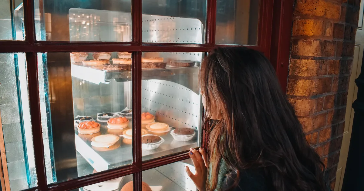 A woman gazes intently at a variety of delicious pastries through a bakery window.