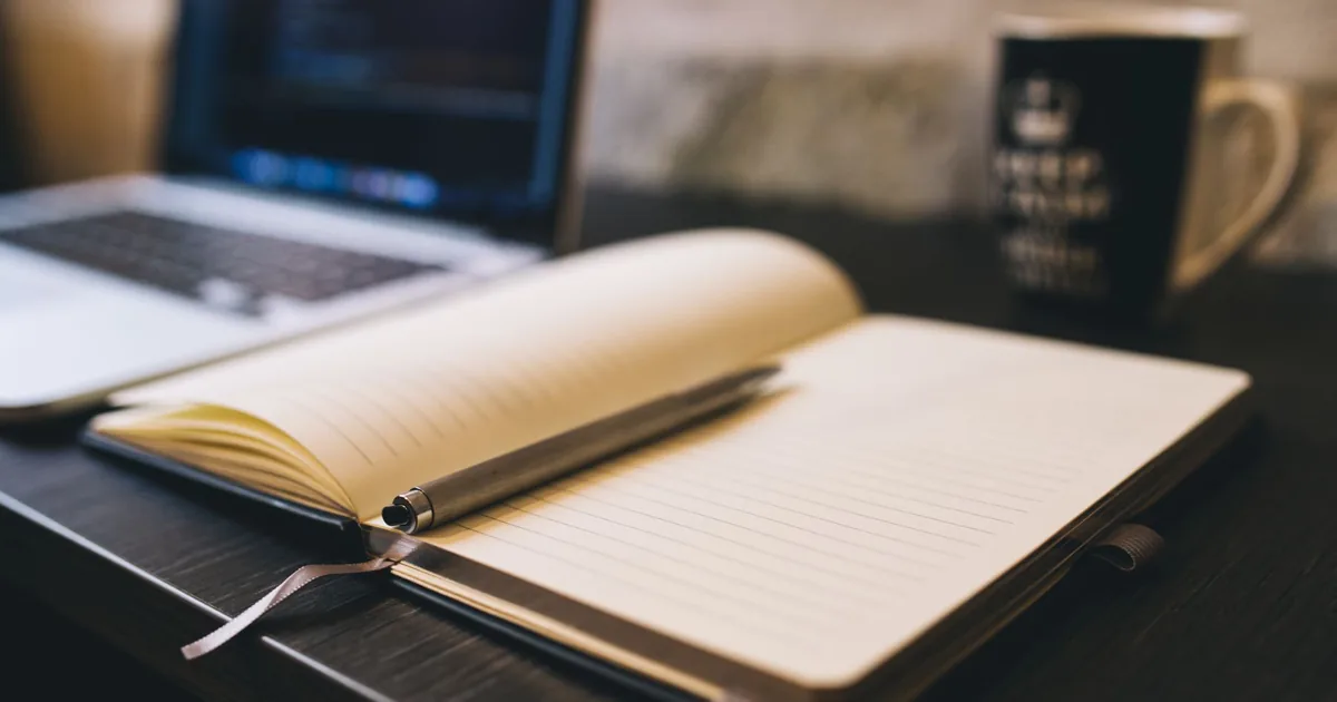 Open notebook with a pen on a wooden desk beside a laptop, ideal for blogging and work-related themes.