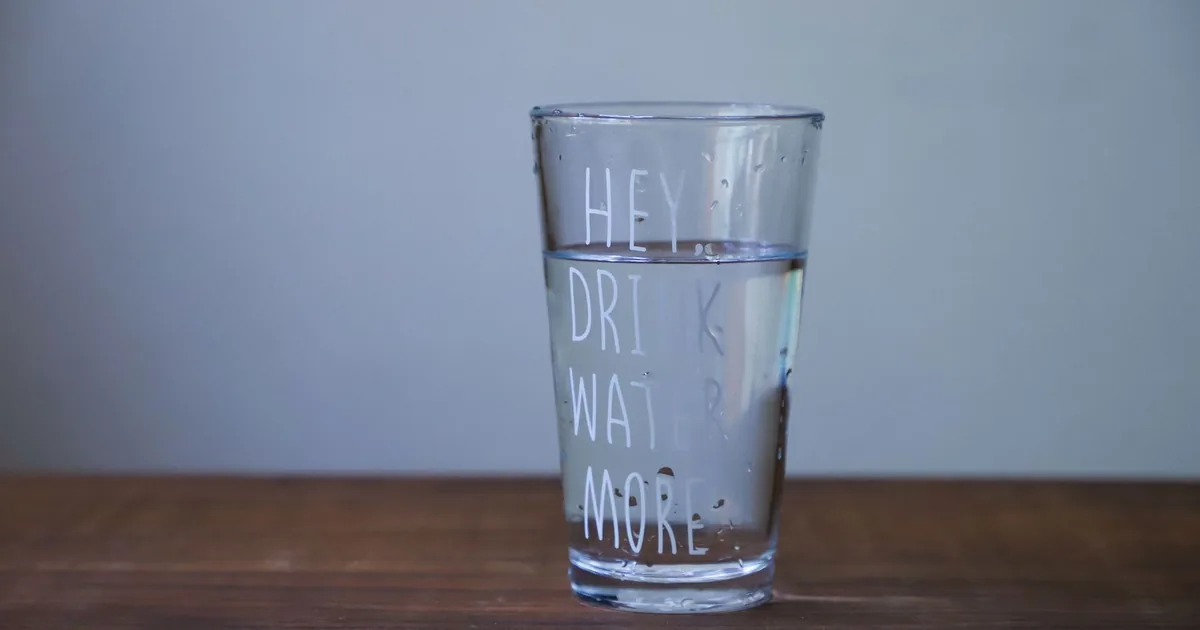 A glass of clear water on a wooden table with the text 'HEY DRINK WATER MORE'.