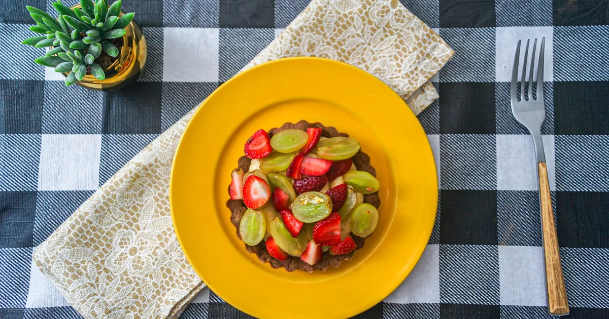 A vibrant fruit tart with strawberries and grapes on a yellow plate, set on a checkered tablecloth for a striking presen