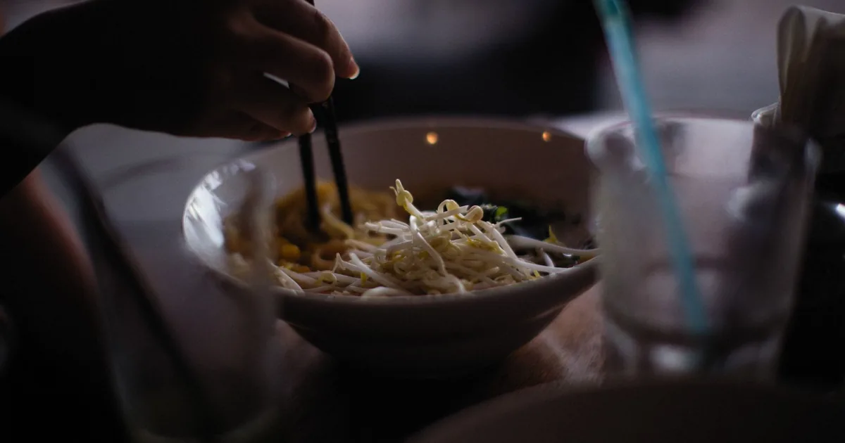 A hand skillfully using chopsticks to eat Asian noodles from a white bowl in dim lighting.