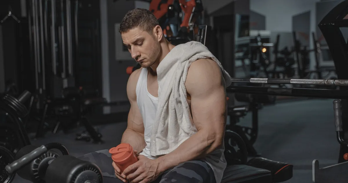Muscular man sits in gym holding protein shaker after a workout, towel on shoulder.