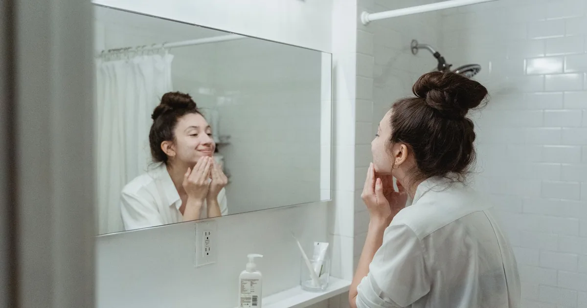 A woman applying skincare cream while looking in the bathroom mirror.