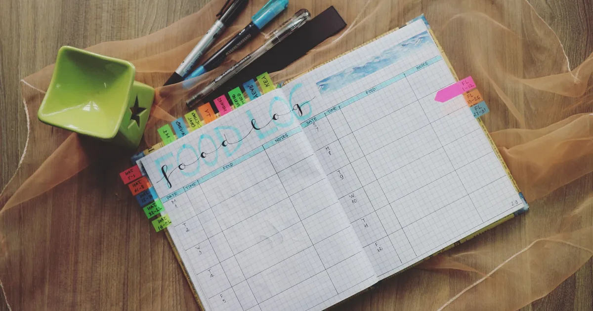 Open food log planner with colorful tabs and pens on a wooden desk, perfect for meal tracking.