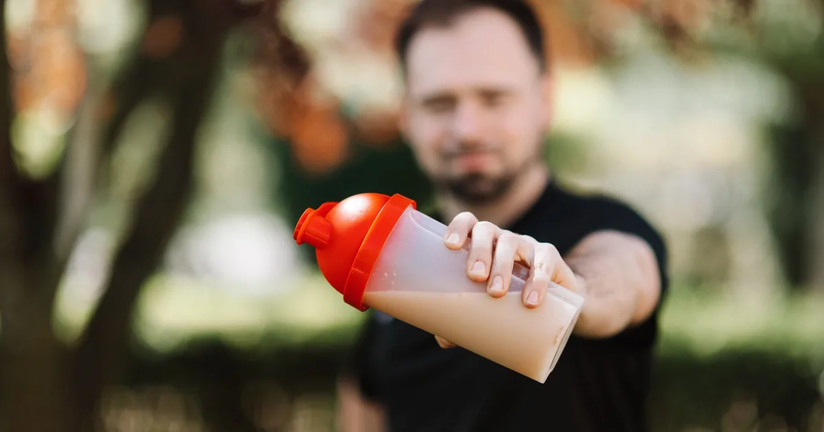 Man presenting a protein shake in a plastic tumbler against a blurred nature background.