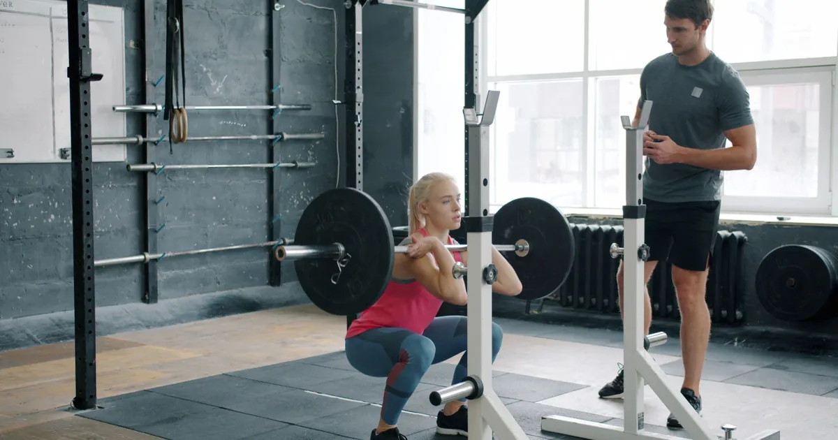 A woman performs a front squat with a barbell, guided by a trainer in a gym.