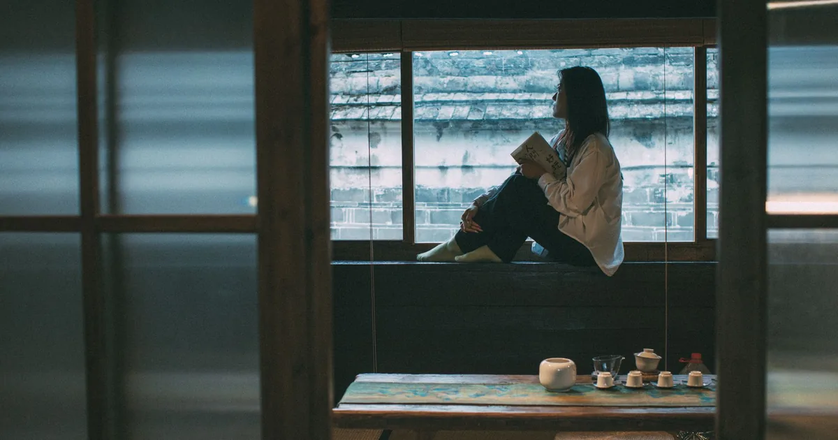 Woman sitting by window in a serene room, enjoying a book and an Asian tea set.