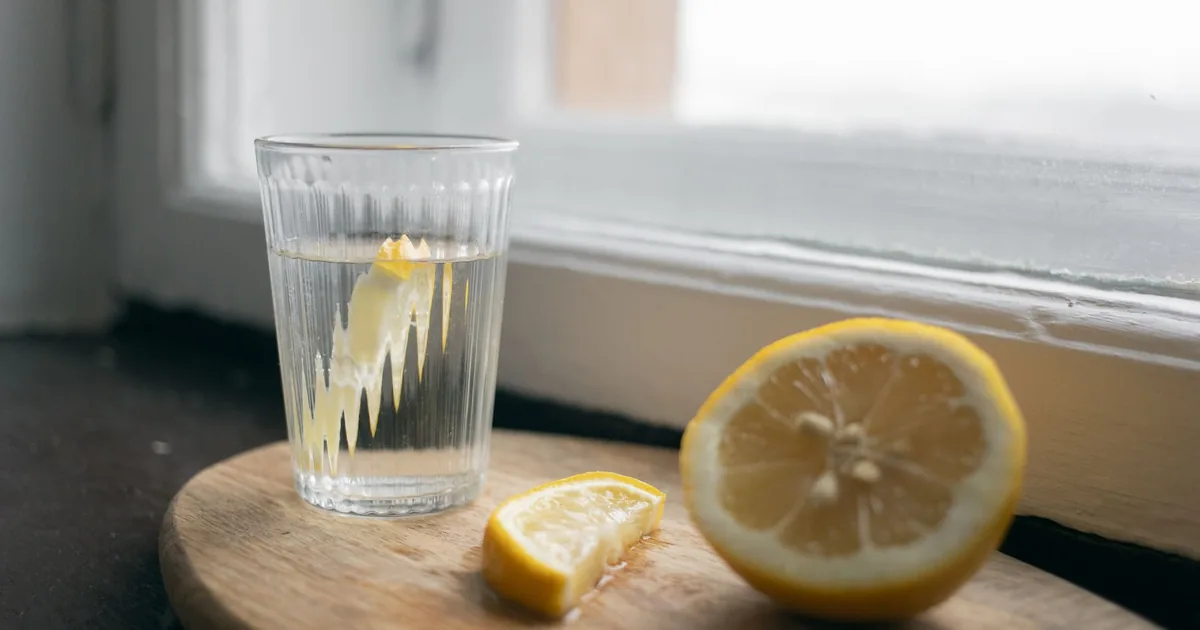 Glass of lemon water with sliced lemons on a wooden board by the window, creating a fresh and cozy vibe.