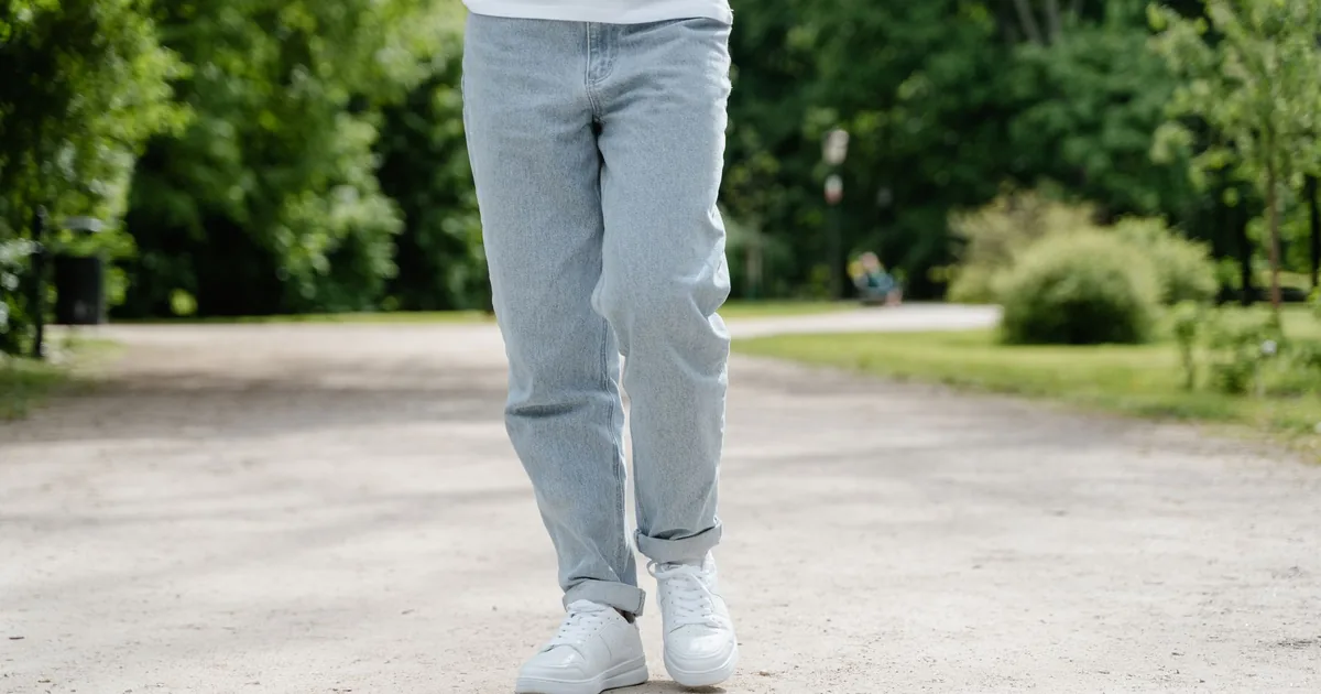 A close-up view of a person walking on a park pathway wearing blue jeans and white sneakers.
