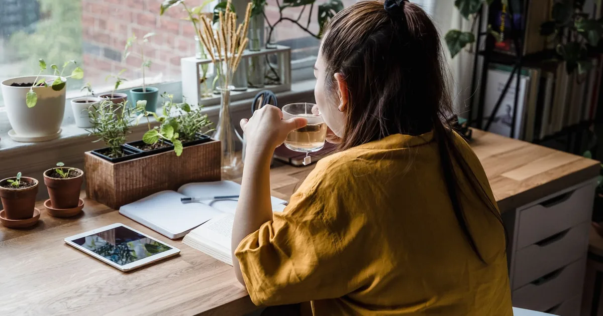 A woman sits at a desk drinking tea, surrounded by plants in a sunlit home office.
